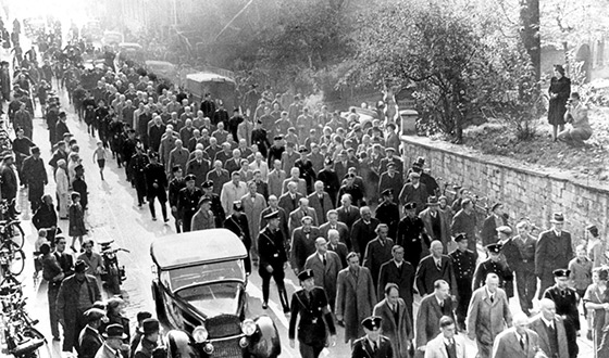 Muchos hombres con sus abrigos recorren una calle en Baden-Baden, vigilados por la SS, hay espectadores. Foto de propaganda de la SS (Derechos de imagen: Archivo Federal)